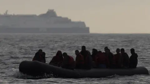 Getty Images A group of about 20 people in an inflatable dinghy on the English Channel. It is dark and only their silhouttes can be seen. In the background a large ferry can be seen