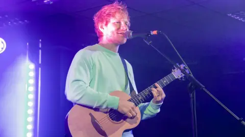 Ella Calver Man with a ginger hair and beard holds a guitar and stands before a microphone. He is strumming the guitar and is wearing a pale green sweatshirt. He is on a stage and is lit from behind in a darkened room.