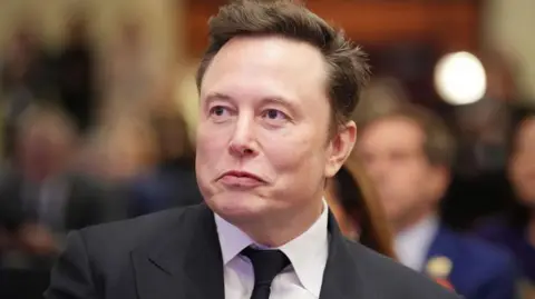 Getty Images Elon Musk wearing a black suit with a white shirt and black tie, with faces of attendees at a House Republicans Conference meeting blurred in the background.