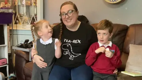 BBC Debbie McCrudden and her two children sitting on a sofa. Conall to the right wearing a red school jumper, and Lilly to the left wearing a grey school dress.