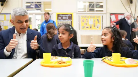 PA Media Mayor of London Sadiq Khan sitting at a lunch table with two school children, doing a thumbs up. He is wearing a dark blue suit and white shirt. The two female pupils have yellow plastic trays with food and a plastic cup in front of them on the table. One of the children is also doing a thumbs up.  