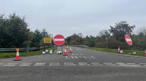 Tony Fisher/BBC One lane of a slip road has been cordoned off by two large no entry signs, red circles with a horizontal white line in the centre. There are also cones cordoning off the slip road in a line from the font of the image around the slip road, until they are out of sight. A yellow sign with black writing reading 'Do not follow sat nav' has also been put on the blocked slip road. On the road, letters written in white paint read 'No Entry'. 