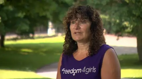 Tina Leslie/Freedom4Girls Tina Leslie, standing in a park wearing a purple singlet top, bearing the logo Freedom4Girls. She is standing in a park with trees in the background.