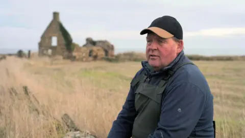 BBC Alan Steven side on to the camera wearing a dark baseball cap with a brown peak, a blue fleece and waterproof dungarees. There is a dilapidated building in the field behind him.