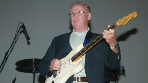 Getty Images Vic Flick performing on stage. He is wearing glasses, a suit jacket and playing a white electric guitar
