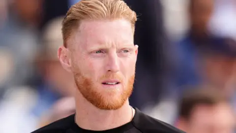 PA Media Close up portrait of Ben Stokes wearing a black top. He has red hair and a beard and is looking slightly to the right. Behind him in the distance is a blurred crowd of spectators.