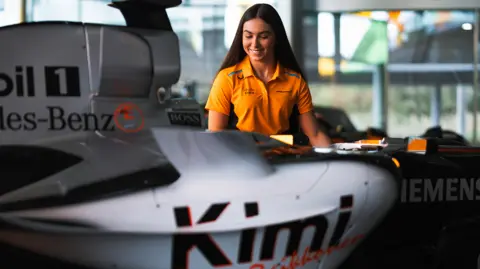 McLaren Racing Ella is sitting next to a McLaren F1 car while she is wearing an orange, team-branded T-shirt 