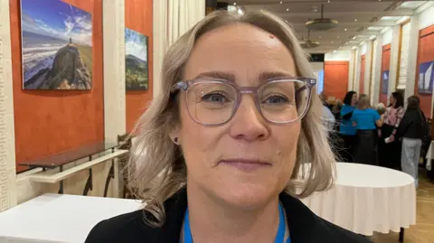 BBC Caroline Campbell - a woman with short, blonde hair wearing grey, square-rimmed glasses. She is smiling at the camera and is standing in the middle of a hall. In the background are some tables with white tablecloths and people talking in the background.