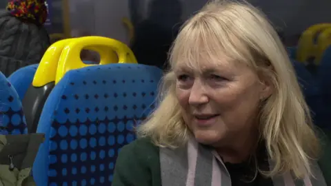 BBC Clare Harland, a woman with blonde hair wearing a green coat, sits on a train