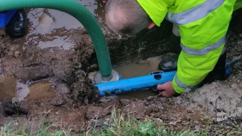 CloudNet A man in a bright yellow jacket leans down as he installs equipment in a muddy trench