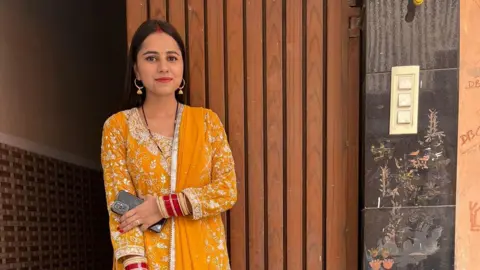 Family A woman in a yellow and white trimmed traditional Indian saree holds her phone in her left hand standing by a wooden doorway.