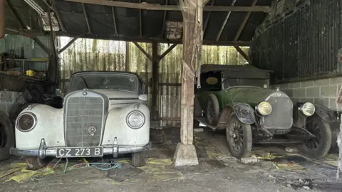 Reeman Dansie Two dusty cars - a white Mercedes from the 1950s and a 1921 green Talbot - pictured with bonnets facing the camera in a dilapidated barn