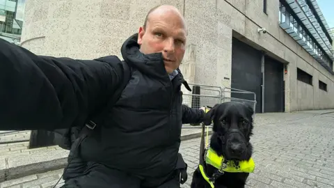 Sean Dilley Sean Dilley is wearing a black jacket as he kneels down to take a selfie with his guide dog Shawn