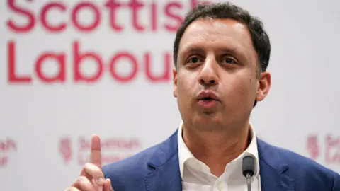 Getty Images Anas Sarwar, with dark hair and wearing a blue suit and white shirt, speaks at a Scottish Labour press conference with his right index finger raised 