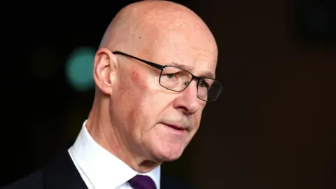 PA Media John Swinney, a bald man wearing glasses and a dark suit with a white shirt, speaks to the media in front of an out of focus black background 