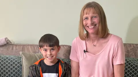 BBC Ruth, wearing a pink top, sits on a sofa next to her eight-year-old son Samuel