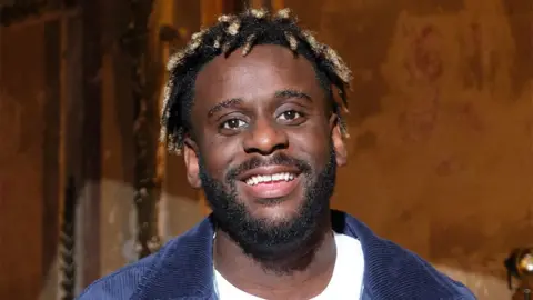 BBC Myles Smith, a man, wearing a white tshirt with a blue denim jacket over the top. He is smiling, with the walls behind him a light brown colour illuminated in some light.