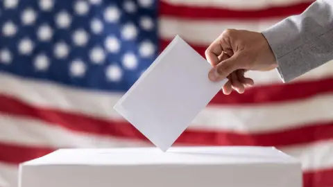 Getty Images Stock image of a ballot being put into a box, with the USA flag behind.