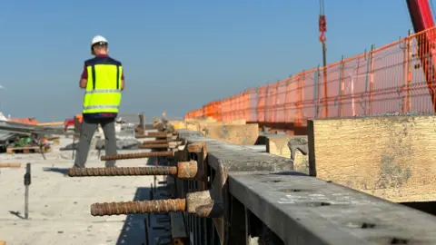 BBC A construction worker in a neon yellow vest and white hard hat stands beside concrete with screws sticking out of it as he works on the new rail project in Lithuania