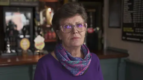 BBC A woman in a purple top, with purple, pink and turquoise snake-skin print scarf. She has short hair and glasses and is looking at the camera with a pensive expression. Behind her a bar and beer taps can be seen.