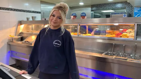 BBC Destiny Harold posing for a photo. She has blonde hair and is behind the counter of a chip shop. There are two men behind her preparing fried food. 