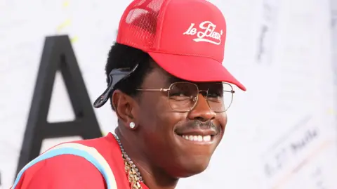 Getty Images Tyler, The Creator wearing a red cap and glasses, smiling.