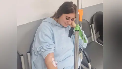 Paige Roberts Paige Roberts sitting down in a hospital chair having IV fluids administered. She is wearing a light blue hospital gown and is looking down at the floor